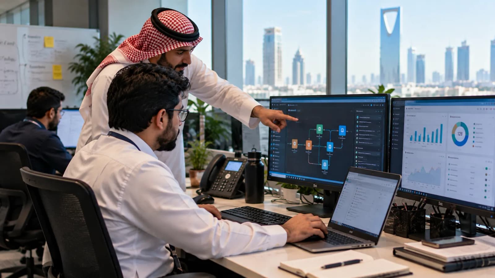 زميل يشير إلى سير عمل على الشاشة بجانب لوحات بيانات الأعمال بينما يعمل موظف على المكتب