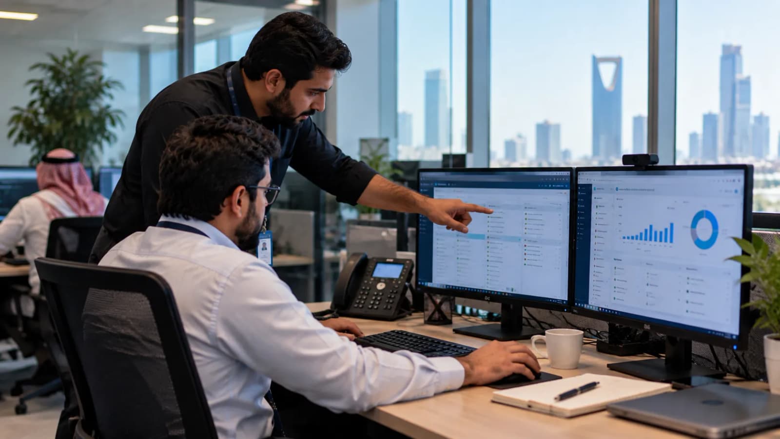 زميلان أمام محطة عمل بشاشتين يراجعان قائمة سجلات العملاء ولوحة مبيعات، وأفق الرياض من نافذة المكتب.