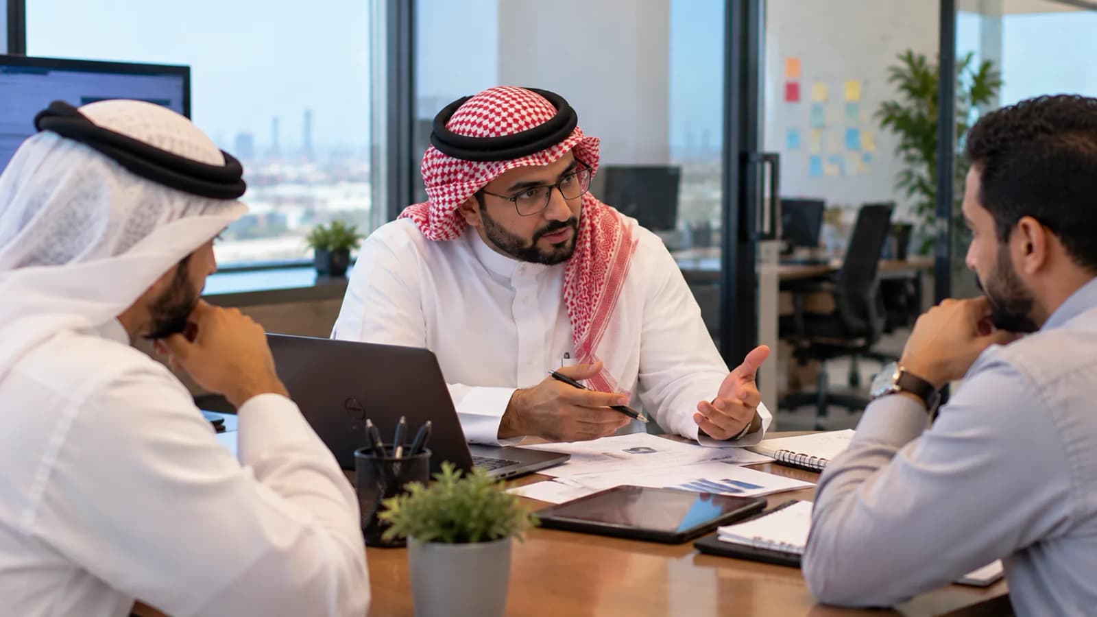 ثلاثة زملاء يتناقشون حول مكتب عليه أوراق وحاسوب محمول وجهاز لوحي، وخلفهم مكتب مفتوح.