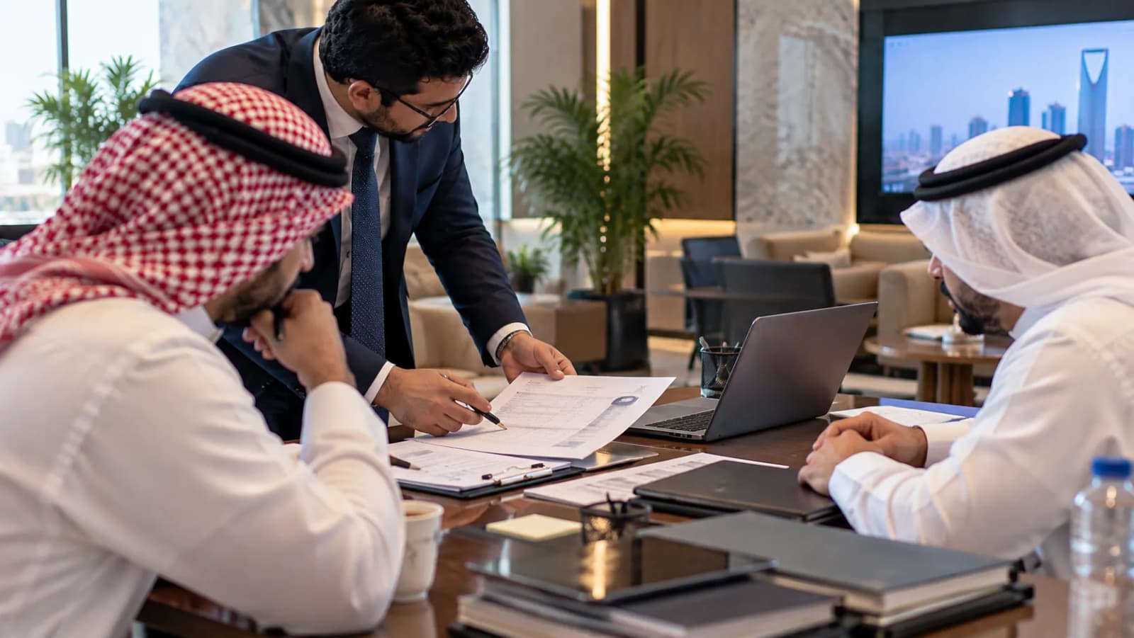 مستشار يقف إلى طاولة اجتماعات ويسلّم نموذج ترخيص مكتملًا إلى عميلين يجلسان أمام حاسوب محمول وملفات.